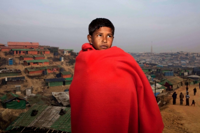 A young boy stands in front of a city with a red blanket wrapped around him