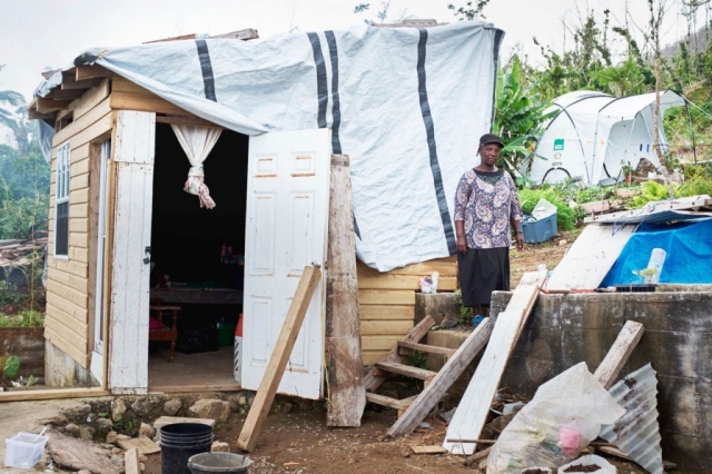 A woman stands in front of her make-shift shelter using a ShelterKit with a ShelterBox tent in the background