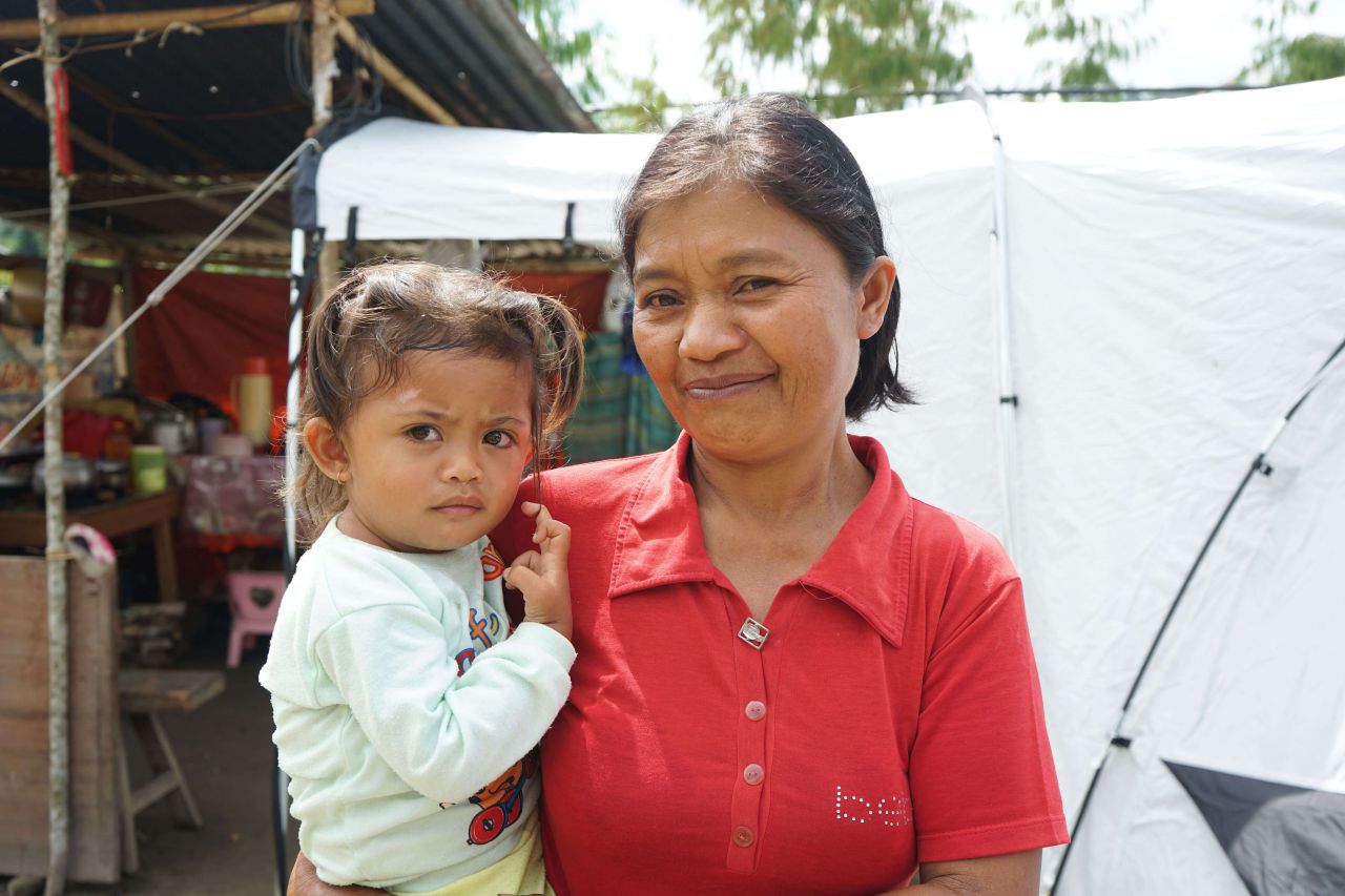 A woman stands with a young child in her arms in front of a ShelterBox tent