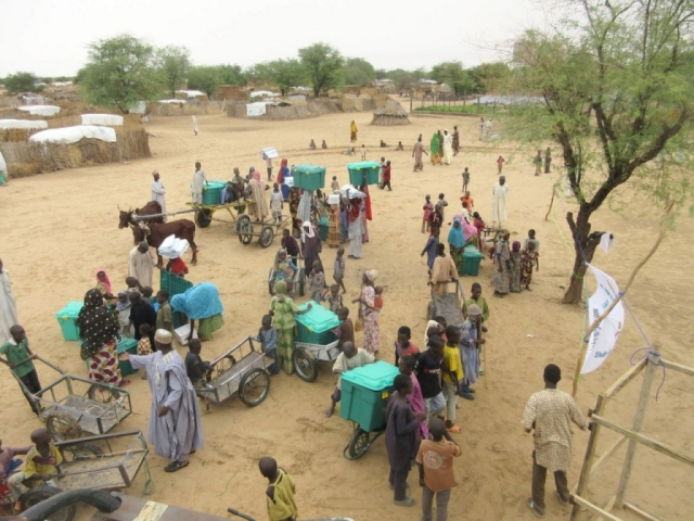A group of people stand in the middle of a large clearing, using bikes and wagons to carry ShelterBoxes