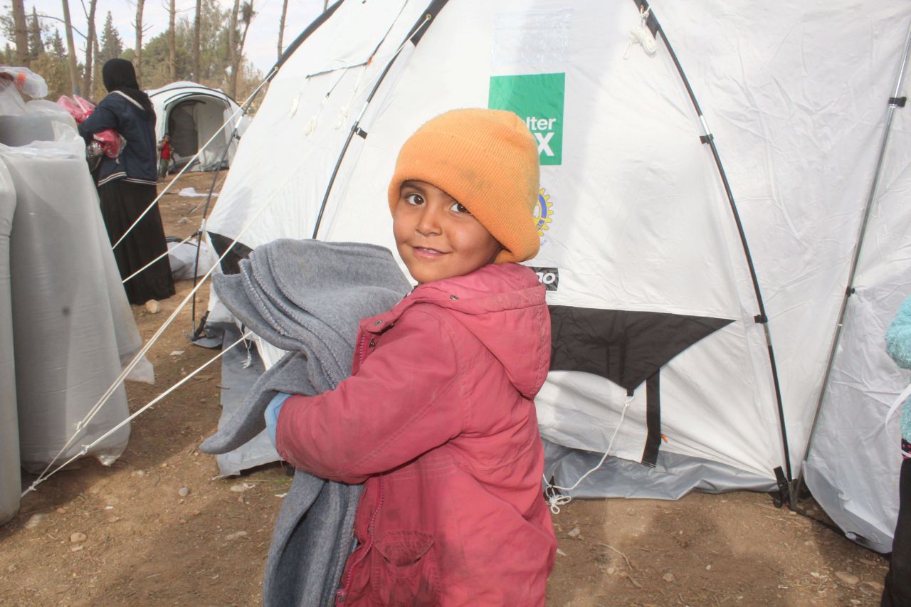 A boy stands holding blankets in front of a ShelterBox tent.