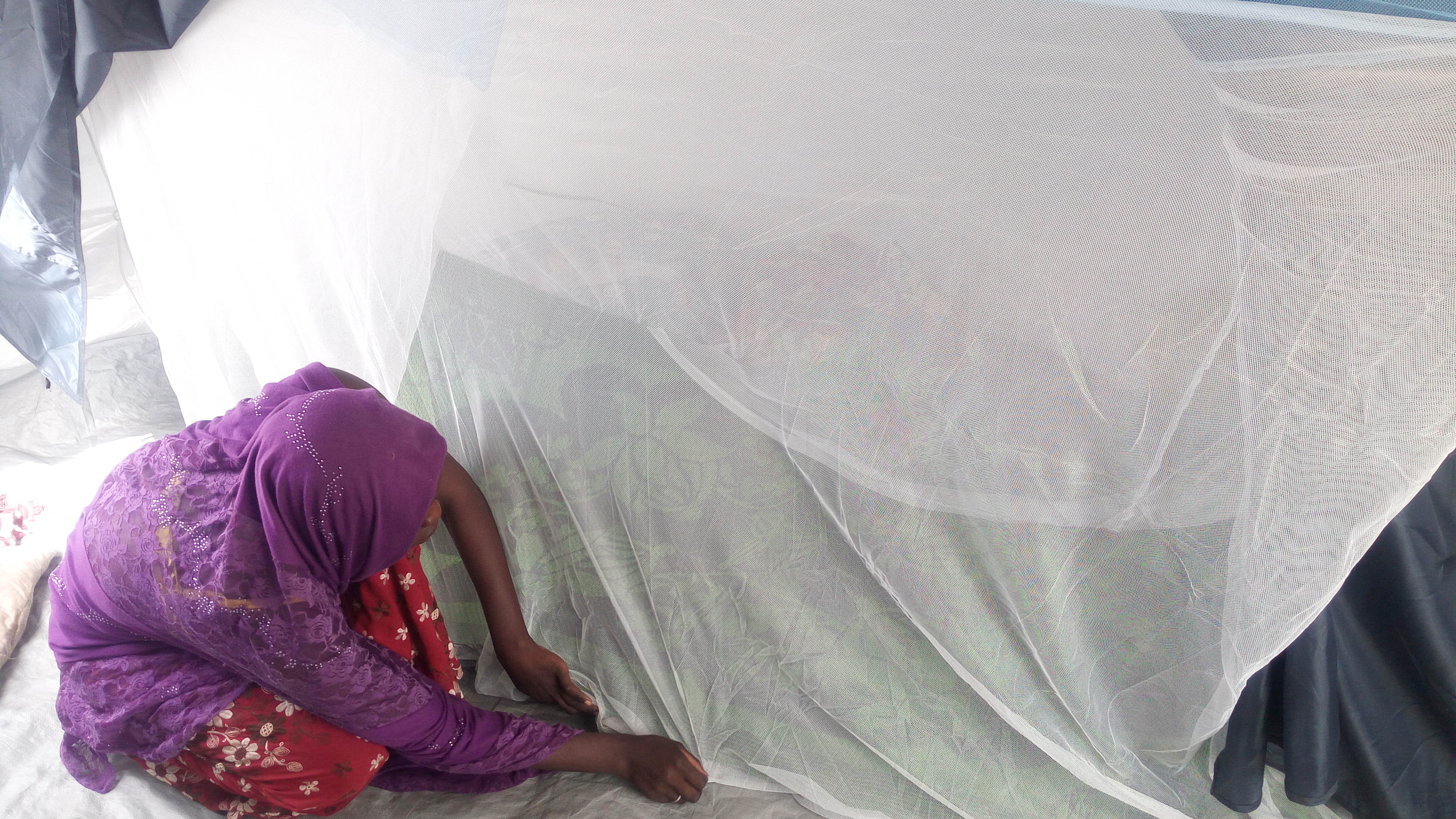 A woman crouches on the floor pulling the edge of a mosquito net to the ground