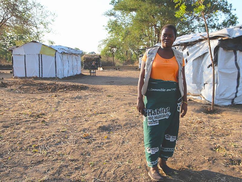 A woman wearing a Habitat for Humanity skirt stands in a field with make-shift shelters around her.