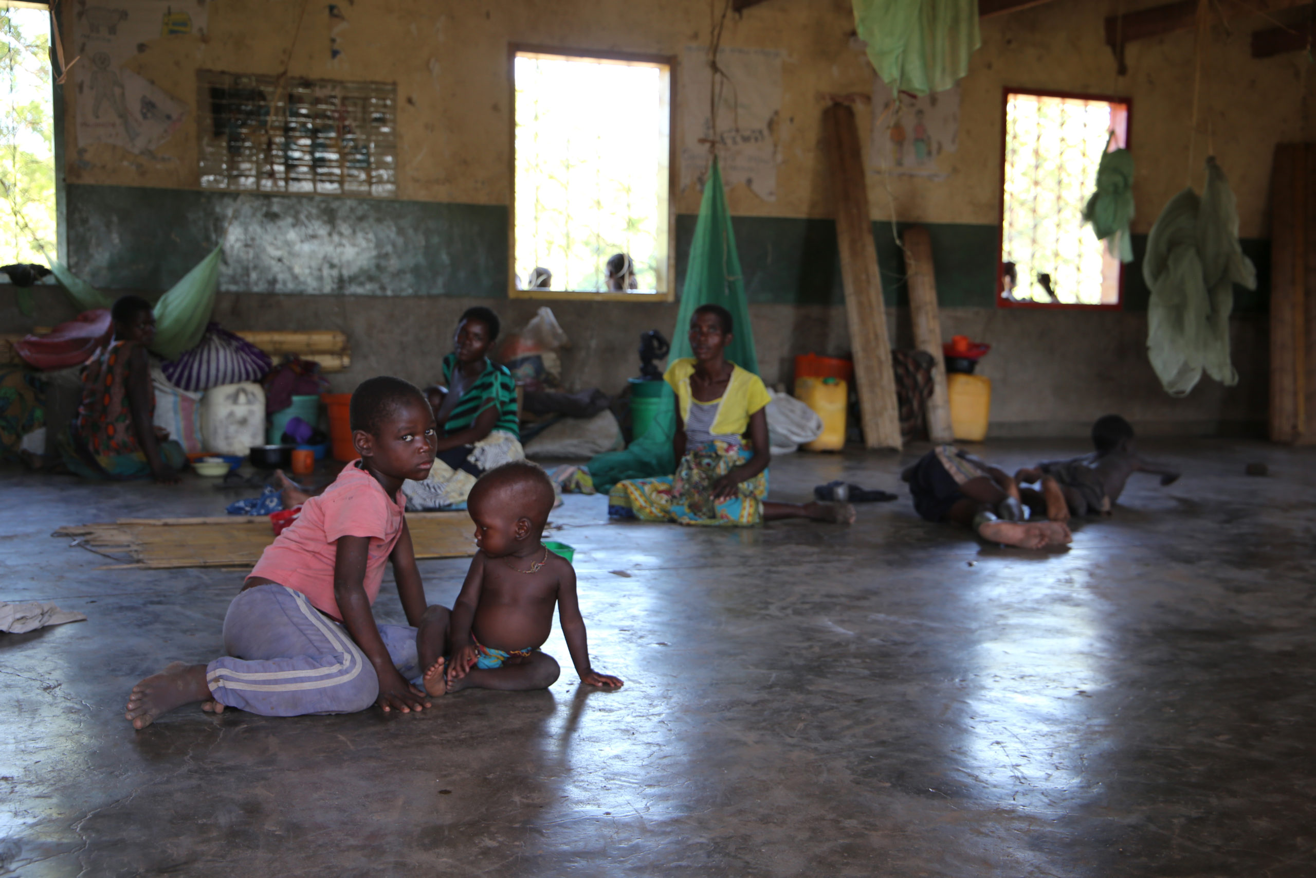 Foreground: two children sit on the concrete ground inside a school being used as an emergency shelter. Adults and other children can be seen in the background