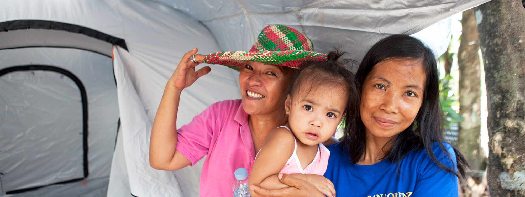 A woman holding a child stands next to another woman in front of a tent. The women are both smiling.