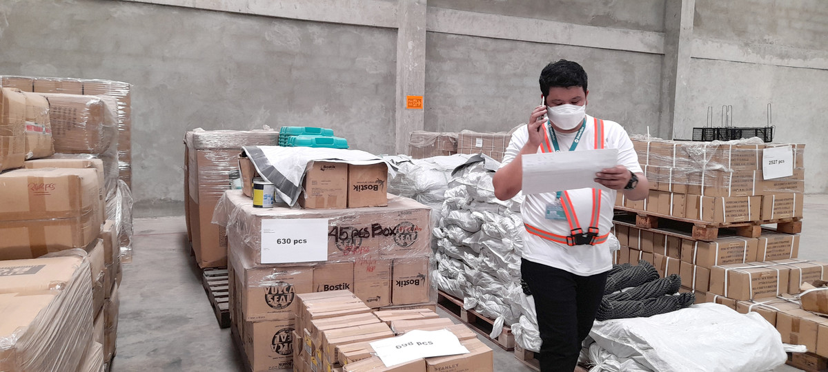 A man wearing a safety vest walking through piles of aid items looking at a sheet of paper