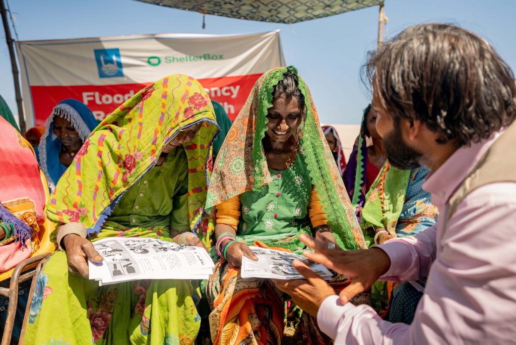 Two people in vibrant yellow and green head scarfs receive training documents from an Islamic Relief support member. They are standing in front of a sign with ShelterBox and Islamic Relief logo's on it. They are smiling and looking down at the training documents.