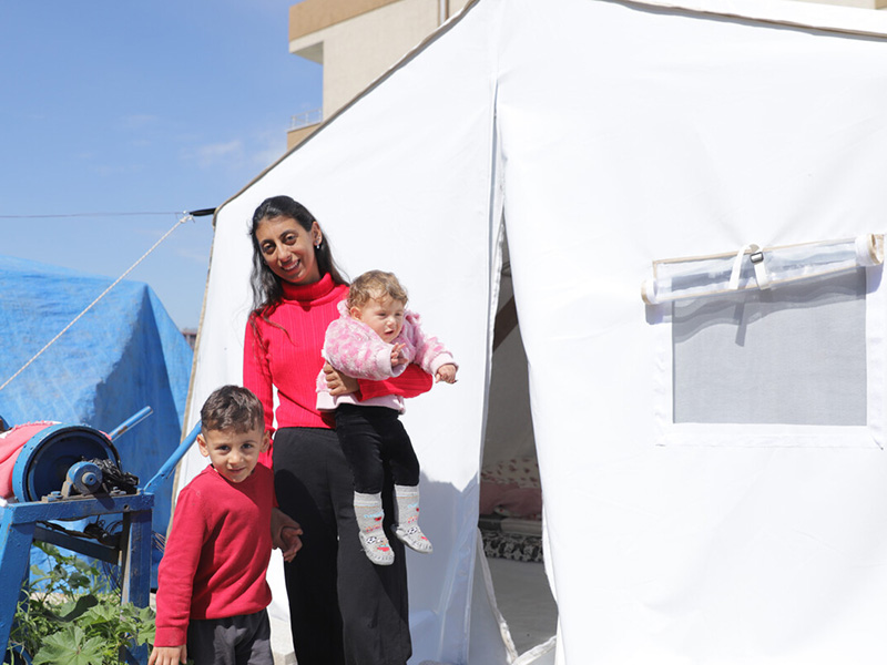A woman stands in front of a tent with her two children